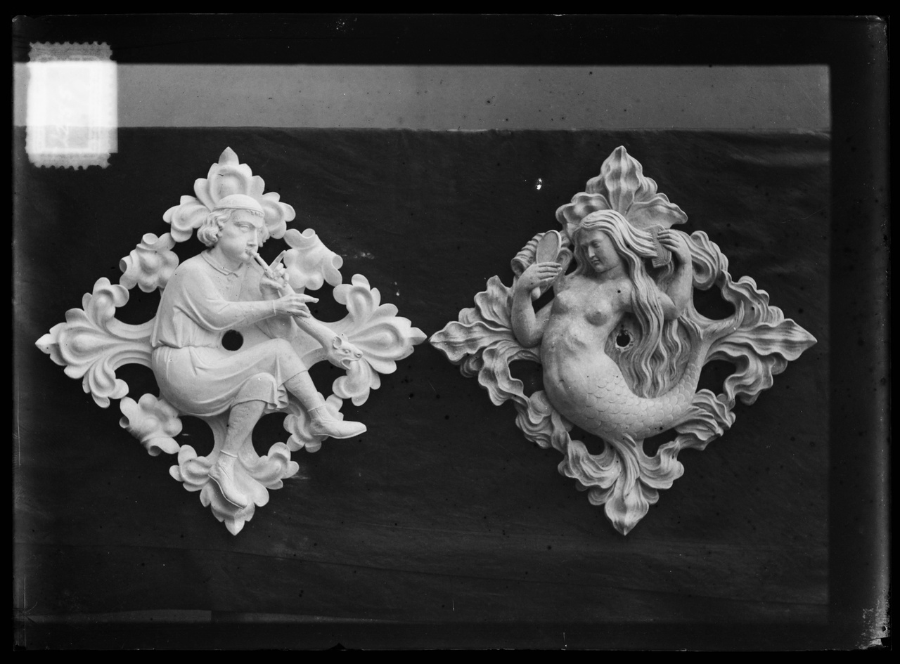 Glasnegatief van foto van producten uit de Cuypers' Kunstwerkplaatsen uit kist met nummer 24: "Twee gipsen muurornamenten met afbeelidingen van een trompetblazer en van een zeemeermin".